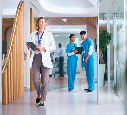 Una dottoressa sorridente attraversa il corridoio di un moderno ospedale tenendo in mano un tablet, mentre due professionisti dell'odontoiatria in camice blu conferiscono sullo sfondo. L'ambiente luminoso e contemporaneo accoglie bambini e famiglie.
