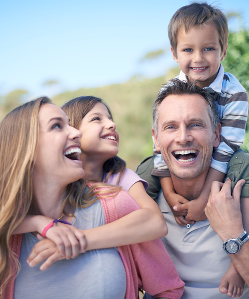 Una famiglia felice di quattro persone sorride all'aperto, mostrando la sua brillante salute dentale. Un uomo porta sulle spalle un bambino, mentre una donna abbraccia una bambina sorridente. I bambini hanno un'aria gioiosa, circondati dal verde sotto un cielo azzurro.