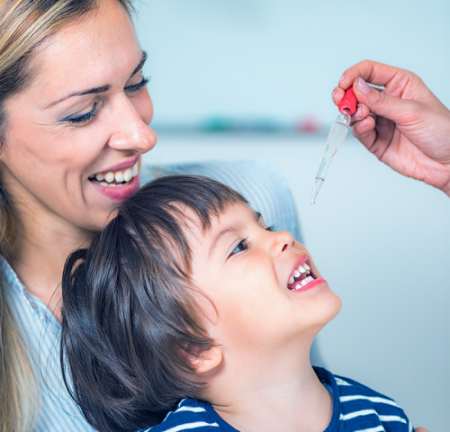 Una donna sorridente tiene in braccio un bambino mentre un'altra persona gli dà medicine o vitamine con un contagocce, promuovendo abitudini sane per i bambini odontoiatrici.