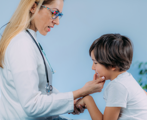 Una dottoressa con gli occhiali esamina la bocca di un ragazzo affidato alle sue cure, mostrando la delicatezza delle cure dentistiche per i bambini. Il ragazzo sembra leggermente a disagio mentre la dottoressa gli tiene il mento e guarda all'interno della bocca.