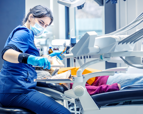 Un dentista che indossa una mascherina e dei guanti lavora su un paziente bambino sdraiato su una poltrona odontoiatrica, utilizzando strumenti dentali in una moderna clinica specializzata nella cura dei bambini.