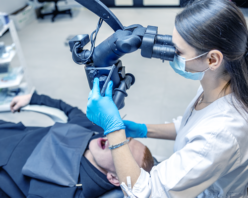 Ortodonzia adulti con un dentista con guanti e mascherina esamina la bocca di un paziente utilizzando un microscopio dentale in clinica. Il piccolo paziente è sdraiato con la bocca aperta e sta ricevendo cure dentistiche specializzate per adulti.