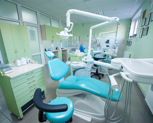 Check up-ortodontico adulti in un moderno ambulatorio dentistico progettato per i bambini, con poltrone dentali color verde acqua, luci a soffitto, armadietti, attrezzature mediche e un interno pulito e luminoso. La stanza ha pavimenti piastrellati e pareti verde pastello con certificati incorniciati.