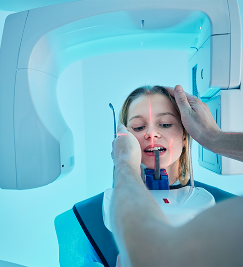 Check up-ortodontico adulti con una donna posizionata in una macchina per radiografie panoramiche dentali, con un tecnico che le regola la testa per prepararla all'imaging. L'ambiente luminoso, con una tonalità blu, sottolinea l'importanza delle cure dentistiche specialistiche per adulti,