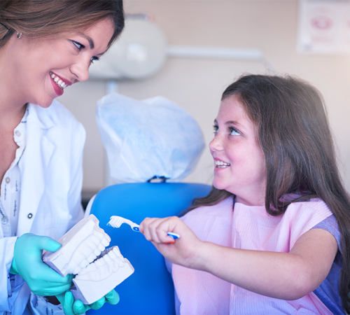 Check-up ortodontico bambini con un dentista con guanti e camice bianco sorride a una bambina sulla poltrona. La sessione di odontoiatria infantile include la ragazza che si esercita a spazzolare i denti su un modello dentale, ed entrambe appaiono felici e impegnate.