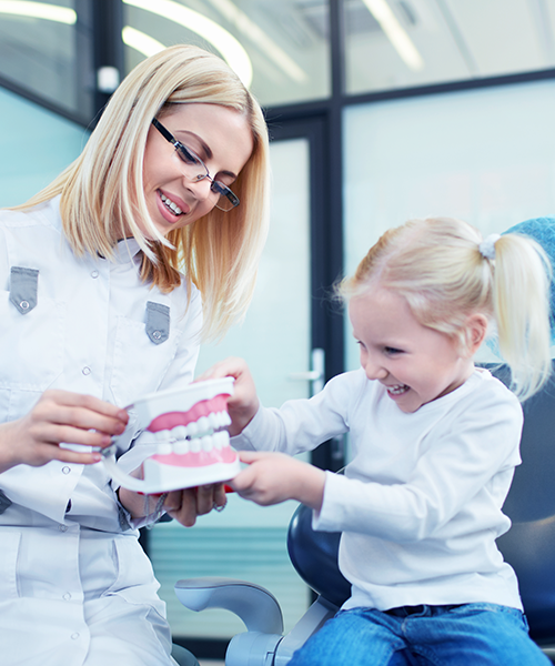 Check-up ortodontico bambini con una dentista mostra un modello dentale a una bambina sorridente in un luminoso studio dentistico. La bambina sembra felice e interessata mentre è seduta sulla poltrona, godendosi la sua visita in questa accogliente clinica dentale per bambini.