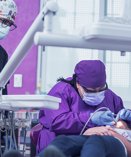 Estrazione dente latte effettuata da un dentista con camice e copricapo viola che esegue un intervento nella bocca di un paziente disteso sulla poltrona, con un’assistente che osserva