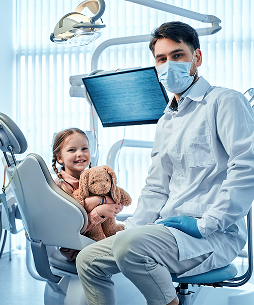 Estrazione dente da latte eseguita da un dentista con camice bianco seduto accanto a una bambina sulla poltrona odontoiatrica che sorride tenendo un peluche, durante una visita per estrazione dente latte in uno studio luminoso