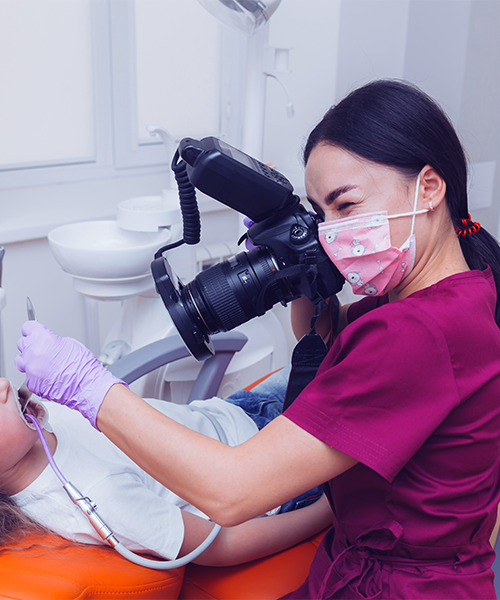 Estrazione dente soprannumerario bambino con una professionista con casacca rosa scuro e mascherina che fotografa la bocca di un bambino sdraiato sulla poltrona odontoiatrica utilizzando una fotocamera professionale con flash, mentre una mano con guanto viola tiene aperta la bocca.
