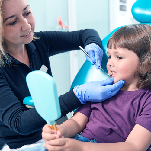 Estrazione dente deciduo durante una visita di controllo, mentre una professionista con guanti blu indica la zona della bocca a una bambina seduta sulla poltrona odontoiatrica che osserva il proprio sorriso in uno specchietto azzurro.