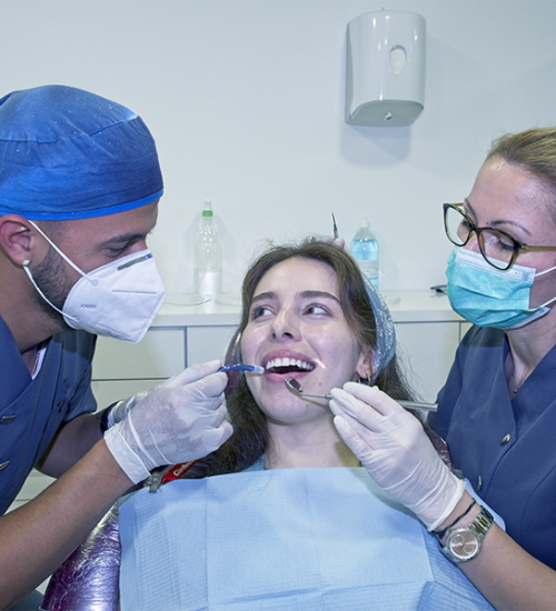 Estrazione dente deciduo durante una visita odontoiatrica, mentre una paziente seduta sulla poltrona sorride e due professionisti con mascherina e guanti utilizzano gli strumenti dentali per esaminare la sua bocca.