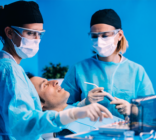 Dental debridement durante una procedura odontoiatrica, mentre due professionisti con camice sterile, mascherina e visiera osservano e preparano gli strumenti accanto a un paziente disteso sulla poltrona in uno studio illuminato da luce blu.