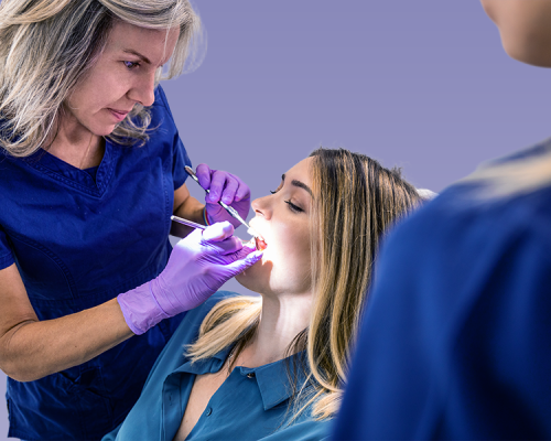 Odontoiatria pediatrica mostrata attraverso una professionista con guanti viola che esamina la bocca di una paziente seduta sulla poltrona odontoiatrica, mentre un’altra persona osserva in primo piano su sfondo lilla.