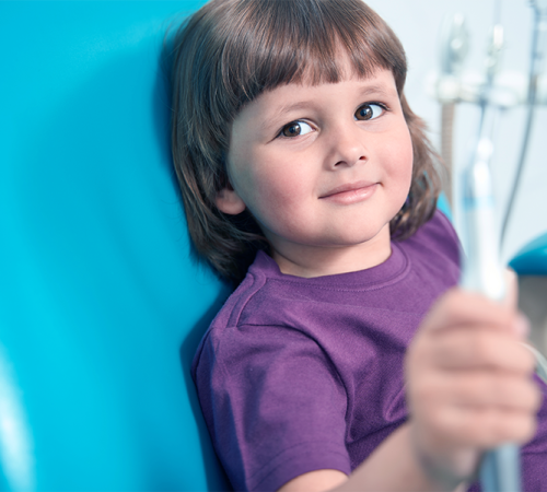 Odontoiatria pediatrica mostrata attraverso il primo piano di un bambino seduto sulla poltrona dentistica, con maglietta viola e sguardo sereno rivolto verso la fotocamera, su sfondo azzurro brillante.
