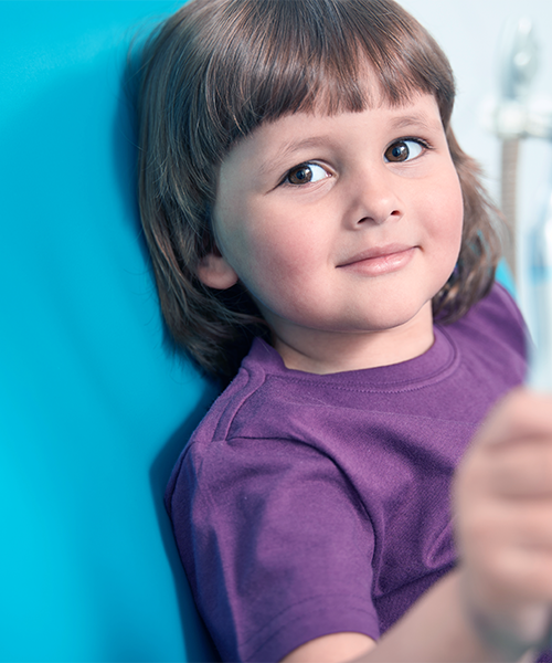 Odontoiatria pediatrica mostrata attraverso il primo piano di un bambino seduto sulla poltrona dentistica, con maglietta viola e sguardo sereno rivolto verso la fotocamera, su sfondo azzurro brillante.