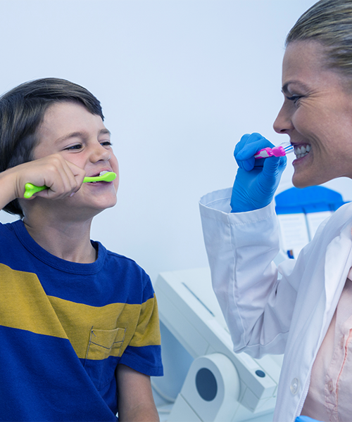 Odontoiatria pediatrica rappresentata da un bambino e una dentista che si spazzolano i denti insieme con spazzolini colorati, mentre la professionista indossa camice bianco e guanti blu e il bambino sorride durante la dimostrazione.