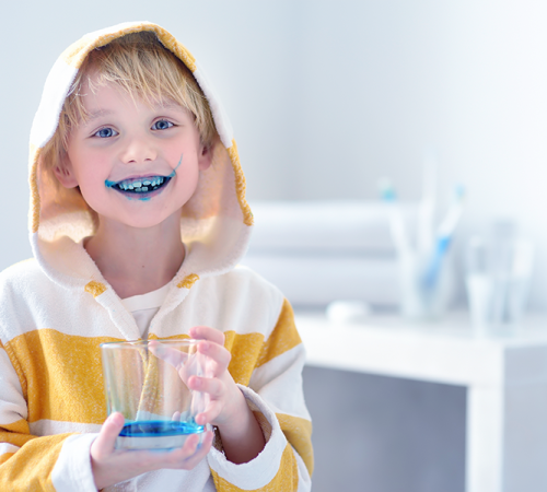 Odontoiatria pediatrica rappresentata da un bambino con la bocca colorata di blu dopo l’uso di un rivelatore di placca, mentre sorride indossando un accappatoio a righe gialle e bianche e tiene in mano un bicchiere con il liquido colorato in un ambiente domestico luminoso.