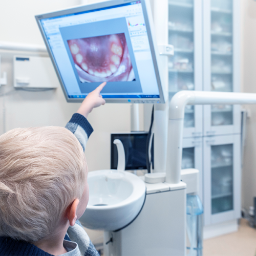 Ricostruzione dente rotto bambino rappresentata da un bambino biondo seduto nello studio dentistico mentre indica con il dito un’immagine ingrandita della propria bocca e dei denti mostrata su un monitor durante il controllo.