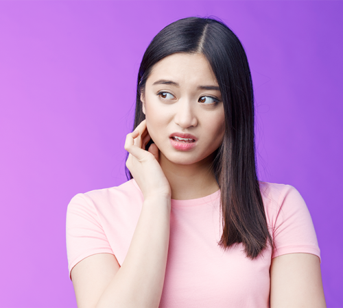 Trauma dentale Ricostruzione dentale rappresentato da una ragazza con espressione preoccupata che si tocca la guancia con una mano, indossando una maglietta rosa su sfondo viola uniforme.