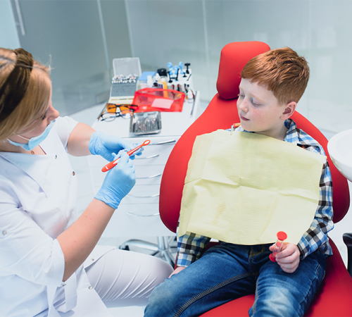 Otturazione dente bambino eseguita da un dentista con camice bianco e guanti azzurri che mostra a un bambino seduto sulla poltrona rossa come utilizzare uno spazzolino, mentre il piccolo indossa un bavaglino giallo e tiene un giocattolo in mano.