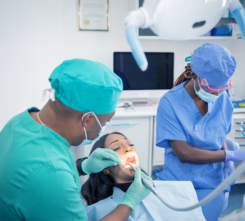 Trauma dentale bambino rappresentato da un dentista con camice e copricapo verde che esegue un intervento nella bocca di una paziente distesa sulla poltrona mentre un’assistente in divisa blu prepara gli strumenti in uno studio odontoiatrico luminoso.