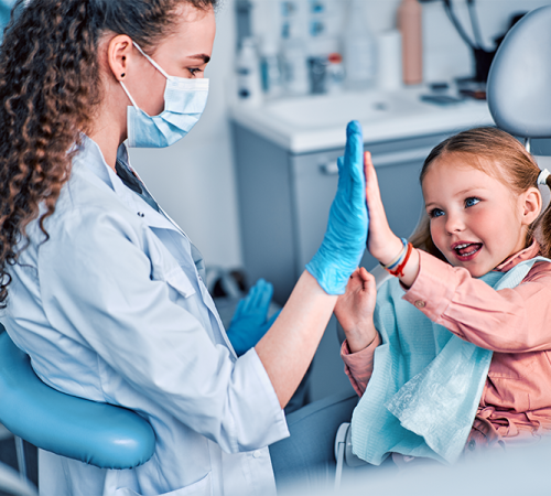 Trauma dentale bambino rappresentato da una dentista con mascherina e guanti blu che dà il cinque a una bambina seduta sulla poltrona odontoiatrica, mentre la piccola sorride con un bavaglino azzurro in uno studio dentistico luminoso.