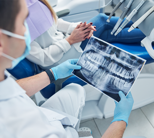 Trauma dentale bambino rappresentato da un dentista con mascherina e guanti blu che osserva e mostra una radiografia panoramica dei denti a una paziente seduta sulla poltrona odontoiatrica con le mani intrecciate in grembo.