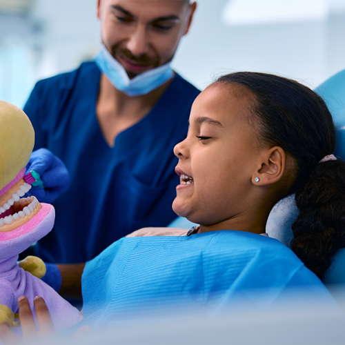Igiene orale bambini rappresentato da una bambina è seduta sulla poltrona odontoiatrica mentre un dentista le mostra come spazzolare correttamente i denti usando un grande modello della bocca, creando un momento educativo e rassicurante.