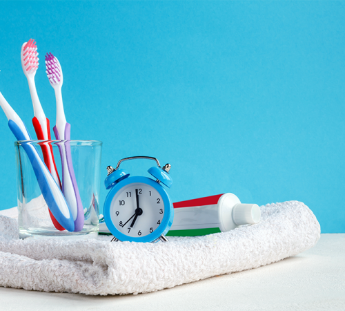 Igiene orale bambini immagine di spazzolini colorati in un bicchiere, un tubetto di dentifricio e una sveglia appoggiati su un asciugamano bianco, simbolo della routine quotidiana di pulizia dei denti per i più piccoli.