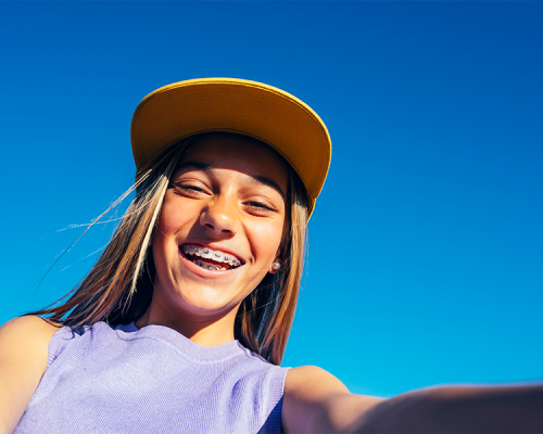 Igiene orale bambini una ragazza sorride all’aperto indossando un cappello giallo mentre mostra i denti in un selfie scattato sotto un cielo blu.