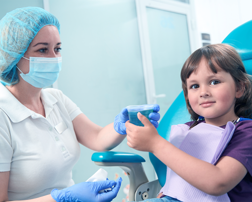 fluoroprofilassi infantile mostra un bambino seduto sulla poltrona dentistica che riceve un bicchierino monouso dalla dentista.
