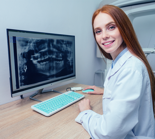 Protesi dentale mostra una donna sorridente in camice bianco seduta alla scrivania, guardando la telecamera, con una radiografia dentale visualizzata sul monitor di un computer di fronte a lei.