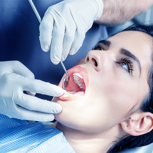 Chirurgia parodontale mostra una donna con i capelli scuri sdraiata su una poltrona odontoiatrica mentre un dentista, indossando dei guanti, le esamina la bocca con degli strumenti dentali. Indossa una pettorina protettiva blu.