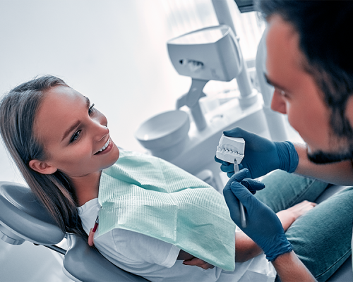 Chirurgia parodontale con un dentista con guanti blu e mostra un modello dentale a una donna sorridente seduta su una poltrona con un bavaglino verde durante una consultazione in una moderna clinica dentale.