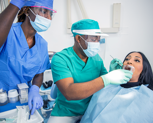 Chirurgia parodontale mostra un dentista e un'assistente dentale che indossano mascherine e camici esaminano la bocca di una paziente in uno studio dentistico illuminato. La paziente è seduta su una poltrona con un bavaglino.