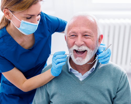 Cure dentali con un dentista con camice blu, guanti e mascherina che esamina la bocca aperta di un uomo anziano sorridente con capelli e barba bianchi in uno studio dentistico.