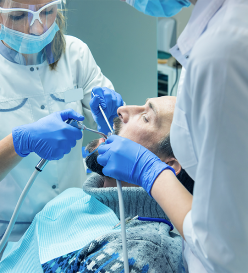 Cure dentali eseguite da due dentisti che indossano mascherine, guanti e occhiali protettivi e che lavorano sui denti di un paziente maschio in uno studio dentistico. Il paziente è reclinato sulla poltrona con un bavaglino dentale, mentre entrambi i dentisti utilizzano strumenti dentali.