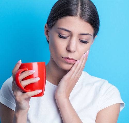 Cure dentali necessarie per una giovane donna con i capelli castani che tiene una tazza rossa in una mano e si tocca la guancia con l'altra, sembrando in preda al dolore o al disagio, su uno sfondo blu.