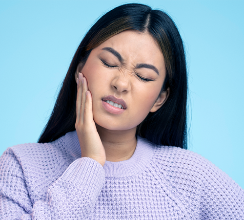 Ipomineralizzazione denti mostra una donna con lunghi capelli scuri che indossa un maglione color lavanda si tiene la guancia e trasale per il dolore, suggerendo un mal di denti o un fastidio alla mascella, su uno sfondo azzurro.