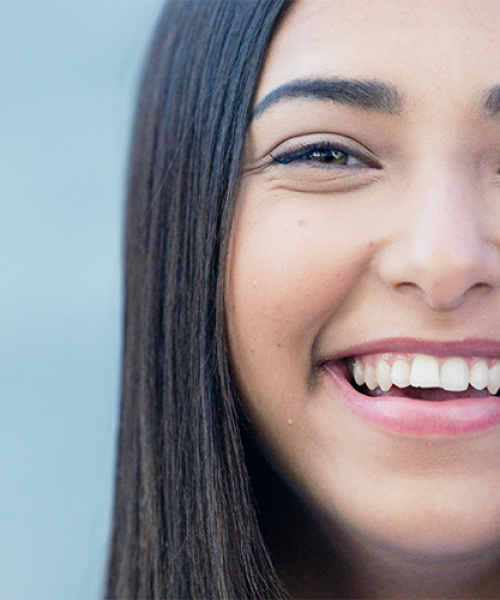Ipomineralizzazione denti bambini rappresentata dal primo piano del sorriso di una ragazza con denti bianchi e regolari, ripresa di profilo mentre sorride su sfondo azzurro sfocato.