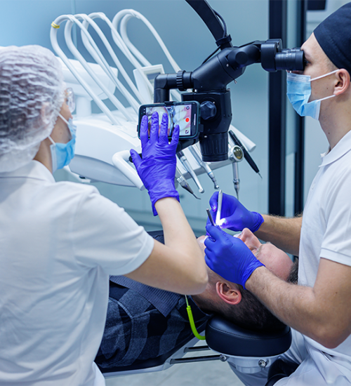Un dentista utilizza un microscopio mentre esamina la bocca di un paziente. Un assistente, che indossa mascherine, guanti e cappucci, tiene in mano uno smartphone, forse per registrare o visualizzare informazioni, in un moderno studio dentistico.