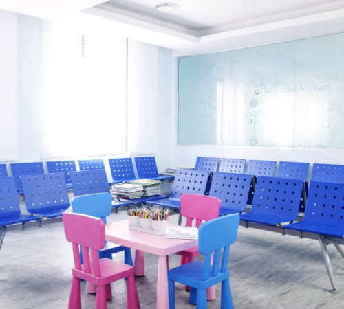 Dental children Milano con la sala d'attesa di Dental Children Milano è illuminata da file di sedie blu vuote, da un tavolo per bambini rosa e bianco con materiale da disegno e da grandi finestre con una parete di vetro smerigliato sullo sfondo.