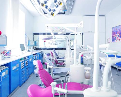 Dental children Milano con il luminoso e moderno studio dentistico a Milano, con diverse poltrone, strumenti e luci in alto. Gli armadietti blu, le pareti bianche e le opere d'arte colorate creano un ambiente pulito e accogliente, perfetto per le visite dentistiche dei bambini.