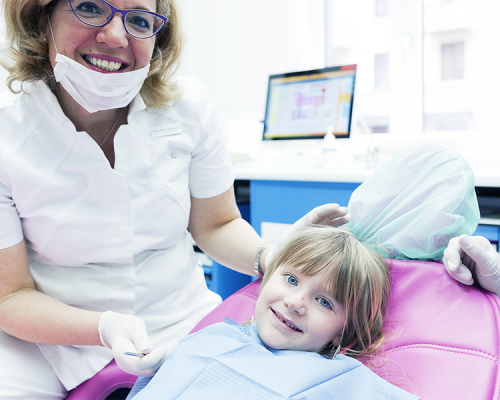 Trauma dentale bambino rappresentato da una dentista con camice bianco, mascherina abbassata e guanti che sorride accanto a una bambina sdraiata sulla poltrona rosa, mentre la piccola osserva la telecamera con un bavaglino azzurro in uno studio odontoiatrico luminoso.