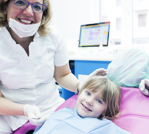 Trauma dentale bambino rappresentato da una dentista con camice bianco, mascherina abbassata e guanti che sorride accanto a una bambina sdraiata sulla poltrona rosa, mentre la piccola osserva la telecamera con un bavaglino azzurro in uno studio odontoiatrico luminoso.