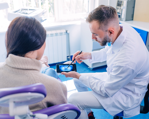 Implantologia dentale mostra un dentista in camice bianco mostra una radiografia dentale su una tavoletta a un paziente seduto con lunghi capelli castani in uno studio dentistico. Il paziente ascolta attentamente mentre il dentista spiega con una penna.