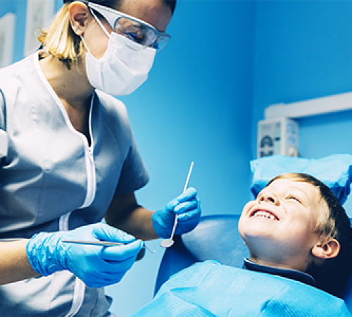 Otturazione dente bambino eseguita da un dentista mentre una professionista con mascherina, occhiali protettivi e guanti blu mostra gli strumenti a un bambino sdraiato sulla poltrona odontoiatrica che sorride, in uno studio illuminato di blu.