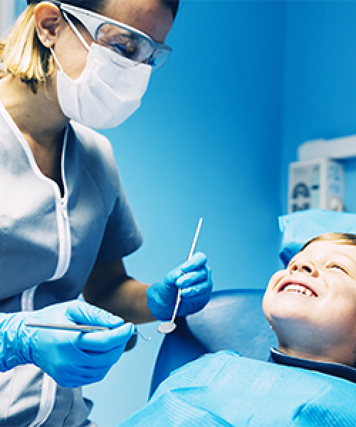 Otturazione dente bambino eseguita da un dentista mentre una professionista con mascherina, occhiali protettivi e guanti blu mostra gli strumenti a un bambino sdraiato sulla poltrona odontoiatrica che sorride, in uno studio illuminato di blu.