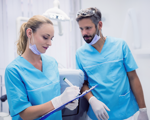 Ipomineralizzazione denti bambini rappresentata da due dentisti in divisa azzurra con mascherine abbassate e guanti, mentre consultano e compilano una cartella clinica su una clip-board all’interno di uno studio odontoiatrico luminoso.
