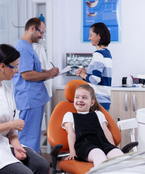 Una ragazza siede sorridente sulla poltrona del dentista mentre un'igienista le parla. Sullo sfondo, due odontoiatri e un'altra donna esaminano le radiografie su uno schermo in una luminosa clinica dentale.